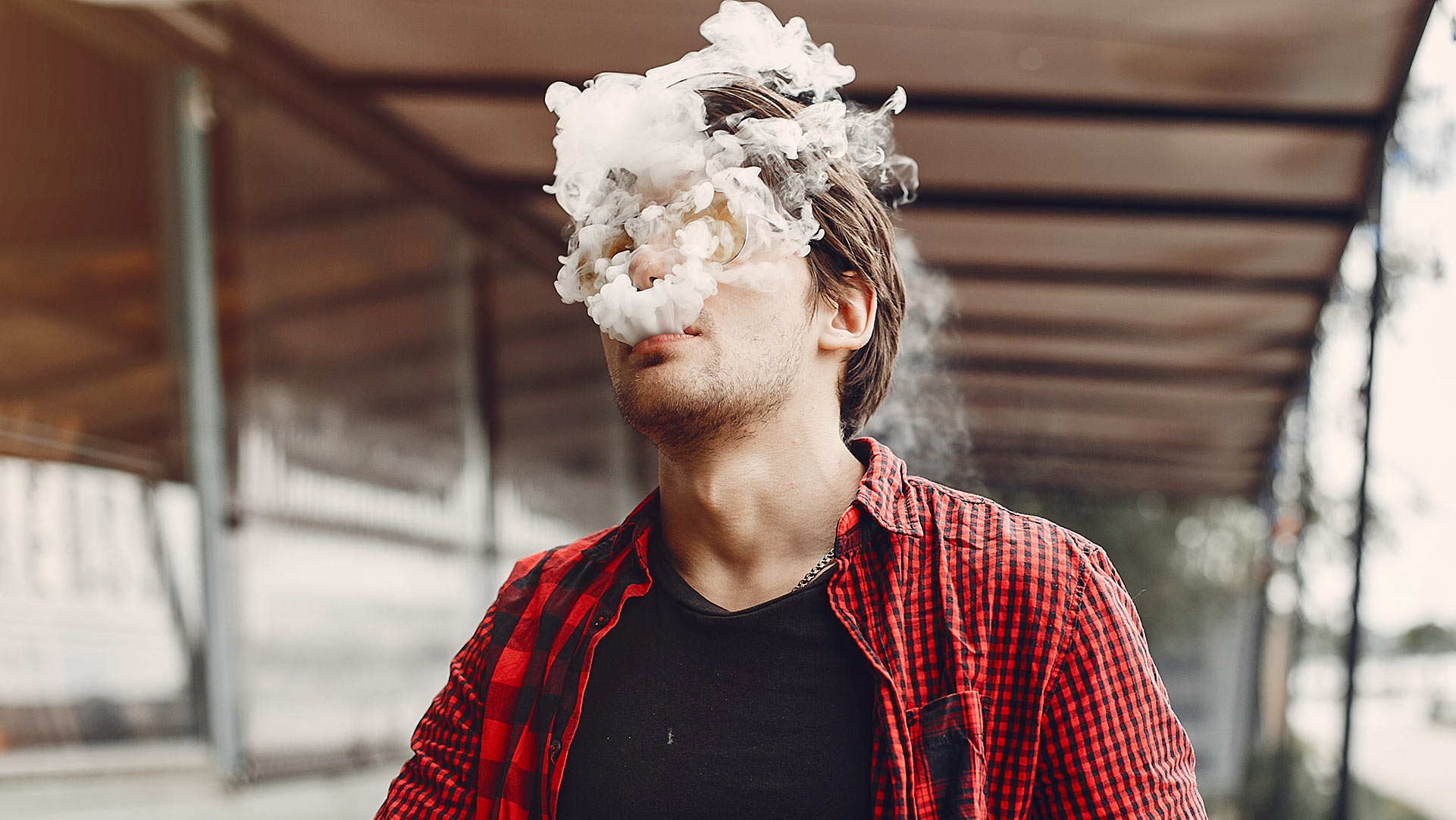 Un giovane con una camicia a quadri rossa e nera che svapa con un fumo denso che gli oscura il volto, su uno sfondo ambiguo dell'aeroporto, pertinente all'argomento del blog "Si può svapare in aeroporto".